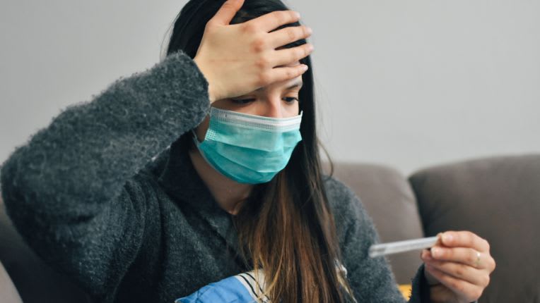 Covid : reprise de l'épidémie à l'automne 2026. Une jeune femme avec la main sur le front, un masque sur le visage, regarde le thermomètre qu'elle tient dans son autre main.