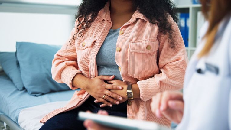 Endométriose : pourquoi cette maladie a longtemps été ignorée ? | ELSAN Actualités. Image d'une jeune femme dont on ne voit pas le visage, qui se tient assise les mains sur le ventre devant une soignante en blouse blanche.