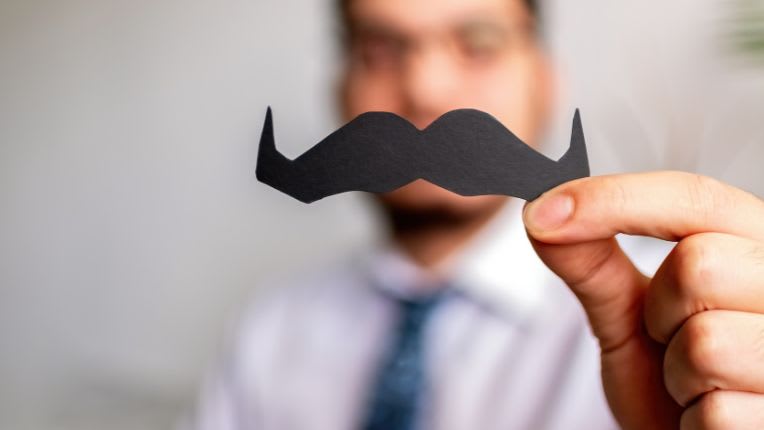 Movember 2025 : agir pour la santé des hommes. ELSAN. Image d'un homme en arrière plan qui présente une moustache noires découpée dans du papier, devant sa lèvre supérieure.