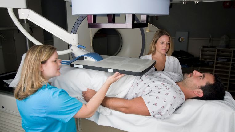 Opération de la prostate par robot à Agen. Image d'un homme allongé préparé par des soignantes pour un séance de radiothérapie.