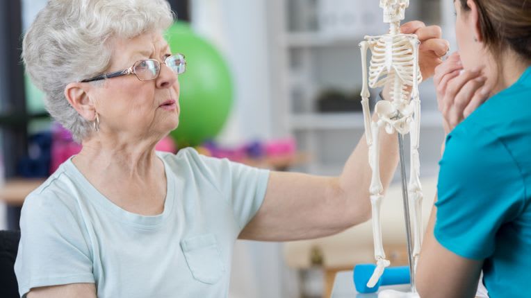 Ostéoporose. Une dame âgées observe un squelette en miniature avec une soignante.
