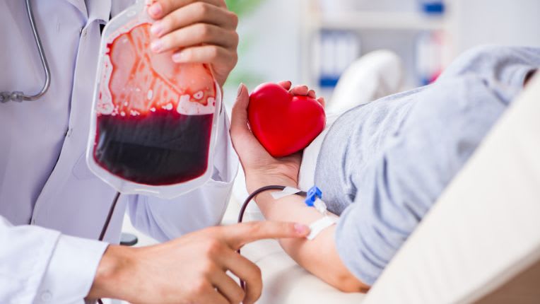 Syndromes myélodysplasiques : image d'un poche de sang et d'un ou d'une patiente allongée, de dos, bénéficiant d'une transfusion sanguine et serrant un coeur rouge en plastique dans la main gauche