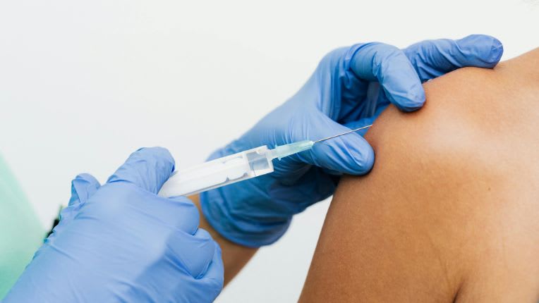 Image en plan serré de deux mains gantées en train d'injecter un vaccin dans l'épaule d'une personnne.