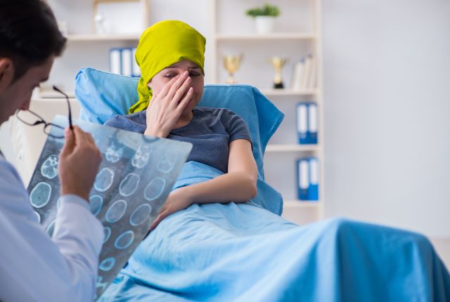 Cancer chez les jeunes et jeunes adultes : augmentation. Un soignant examinant des clichés d'imagerie médicale devant une jeune femme avec un foulard jaune sur la tête, alitée dans une chambre d'hôpital et en pleurs.