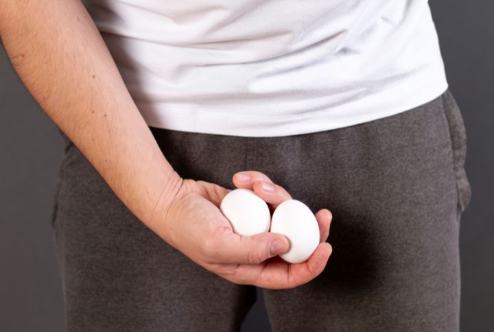 Dépistage du cancer des testicules : un homme soupèse deux œufs devant sa braguette, simulant une autopalpation.