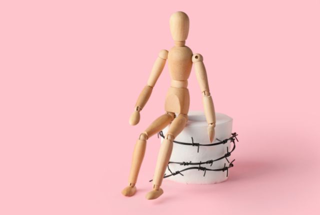 Bonhomme de bois assis sur un siège cerclé de barbelé pour illustrer la définition d'une fissure anale. Une fissure anale est une plaie se situant sur la marge anale. 