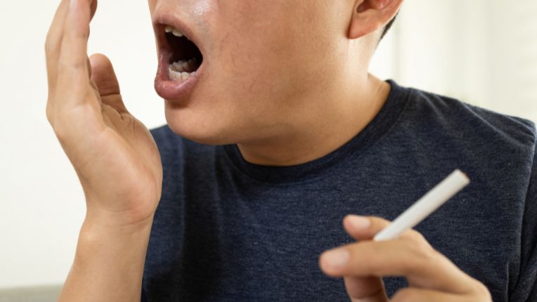 Mauvaise haleine ou halitose : un homme, cigarette à la main, souffle dans le creux de son autre main.