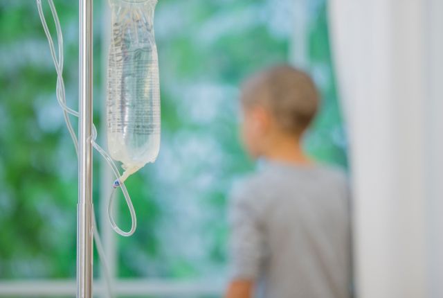 Quelles sont les causes de la leucémie lymphoblastique aiguë ? Image d'une poche de perfusion avec à l'arrière plan, en flou, un jeune enfant, le crâne presque nu, regardant par la fenêtre.