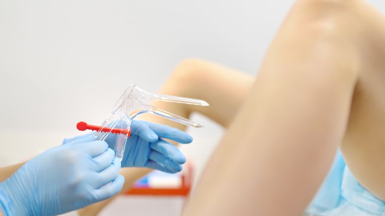 Frottis. Image des mains d'un soignant gantée tenant un spéculum transparent devant les jambes écartées d'une patiente.