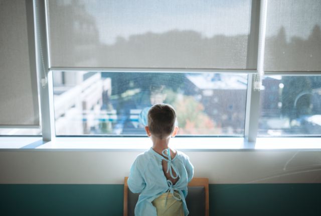 Sarcome des tissus mous : définition, symptômes, causes. Jeune enfant de dos, portant une blouse bleue, juché sur une chaise, regarde l'extérieur par la fenêtre de sa chambre d'hôpital.