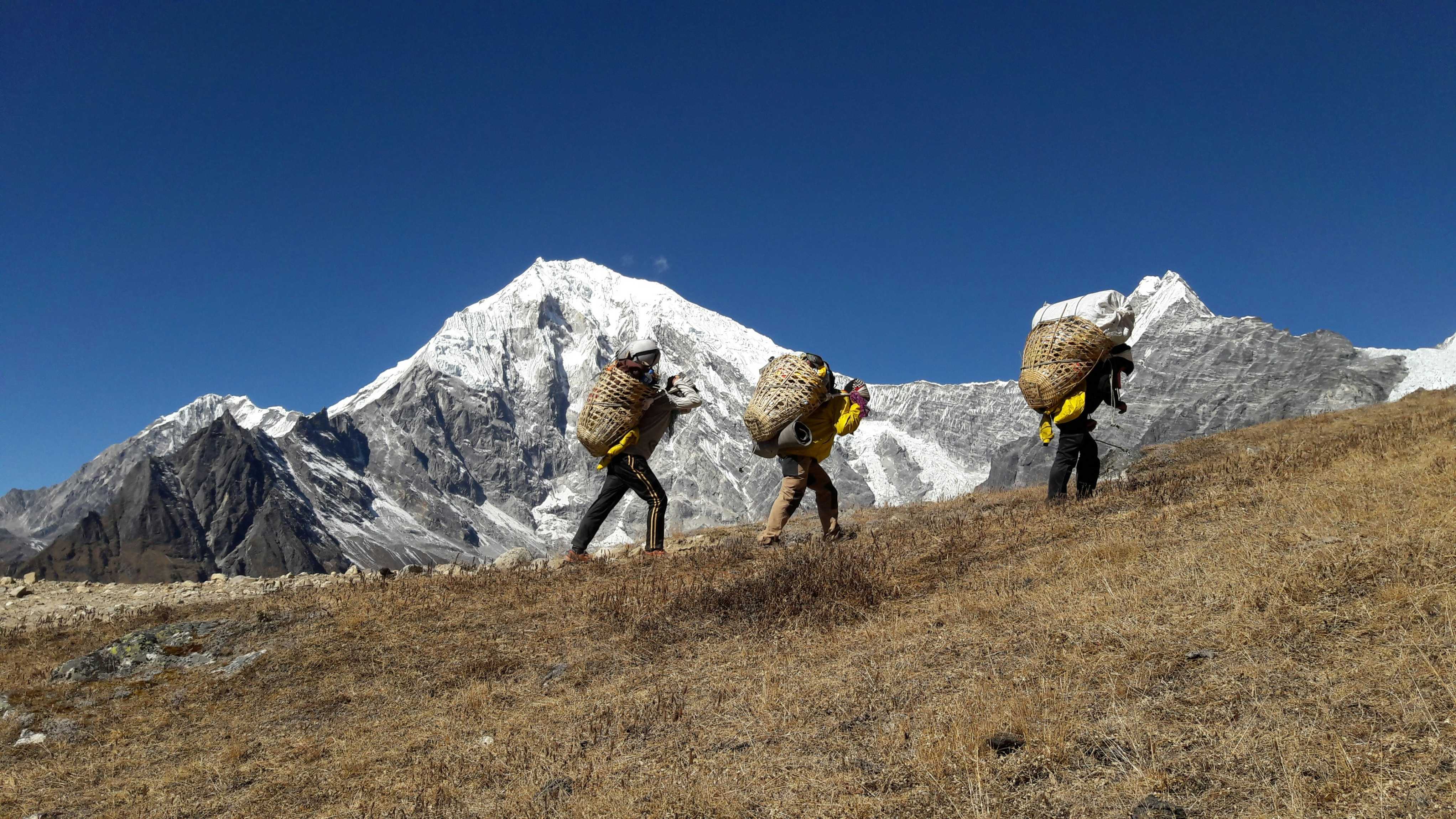 Local Porter Carrying Bamboo Doko Basket with Langtang Lirung Peak in the Horizon A local Nepali porter walking along a hillside trail, carrying a traditional bamboo doko basket, with the towering snow-capped peak of Langtang Lirung at 7,227 meters dramatically visible in the background, Langtang Valley, Nepal.