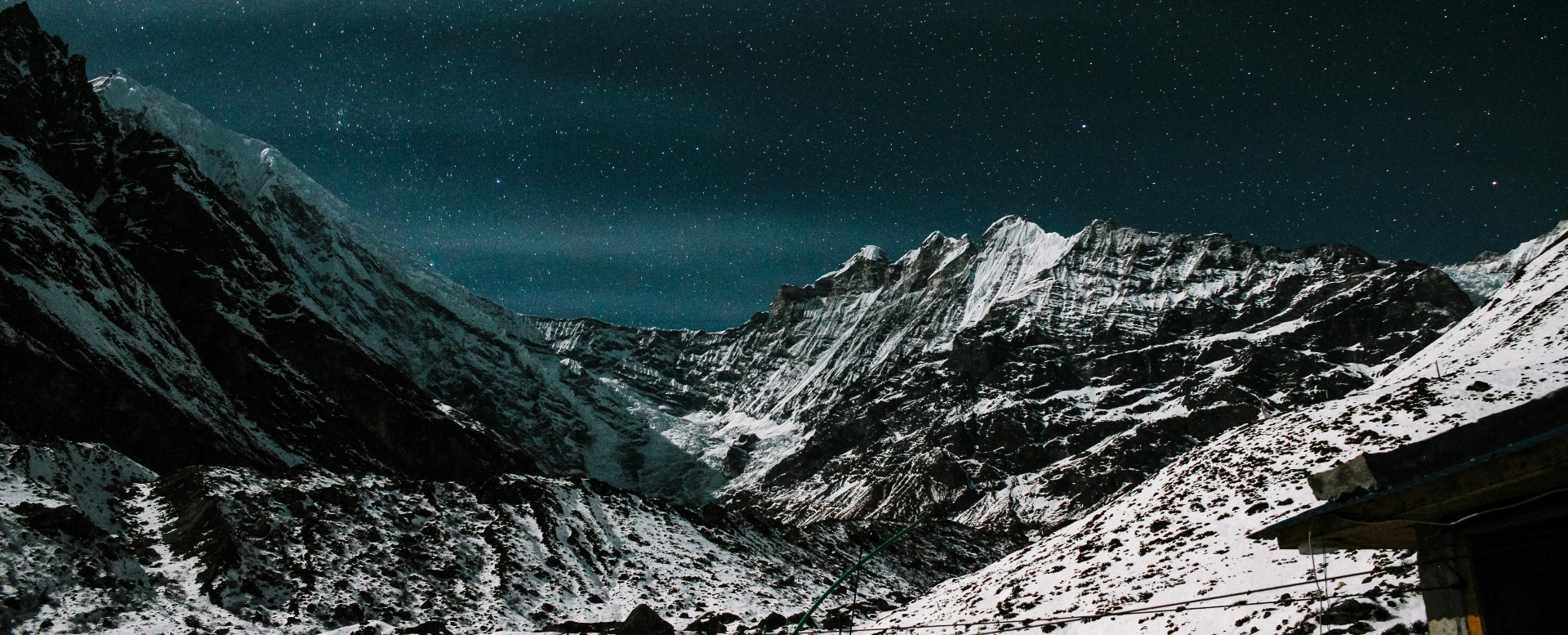 Starlit Serenity Above Kyanjin Gompa - Nocturnal Himalayan Majesty, Langtang, Nepal A breathtaking nighttime photograph of the snow-capped Himalayan peaks in the Langtang region of Nepal, illuminated beneath a vast, star-studded sky. A small traditional teahouse or mountain hut is partially visible in the lower right corner, evoking the warmth and shelter of high-altitude trekking settlements such as Kyanjin Gompa, set against the serene and awe-inspiring nocturnal alpine landscape.