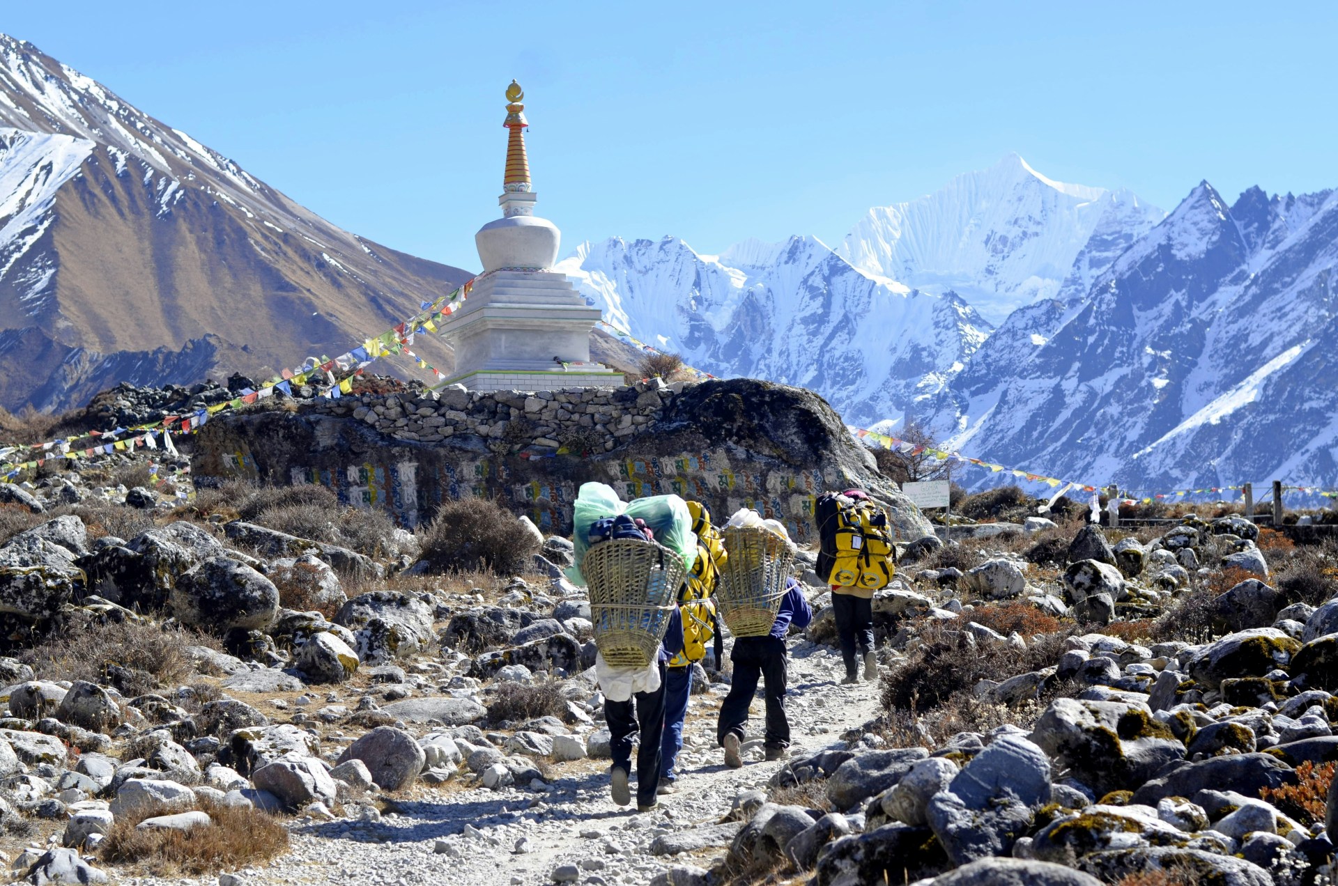a chorten adorned with traditional prayer flags, situated in the Langtang National Park. Porters are visible carrying large, heavy loads in wicker baskets along the rocky trail toward the snowy peaks.This location is a popular trekking route known for spectacular views of the Langtang Lirung mountain range.