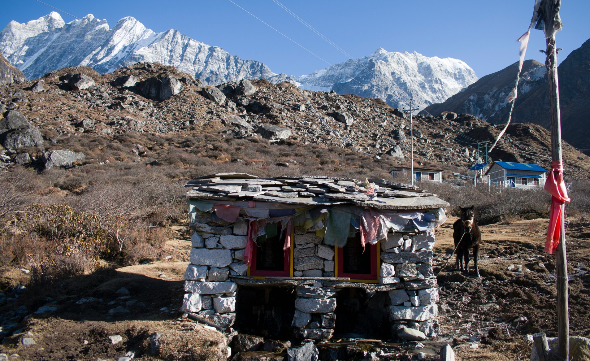 traditional stone structure adorned with colorful prayer flags. a Langtang Valley Trek in Nepal, likely featuring a traditional stone structure adorned with colorful prayer flags.The landscape is characterized by high-altitude, rocky terrain and distant snow-capped Himalayan peaks.