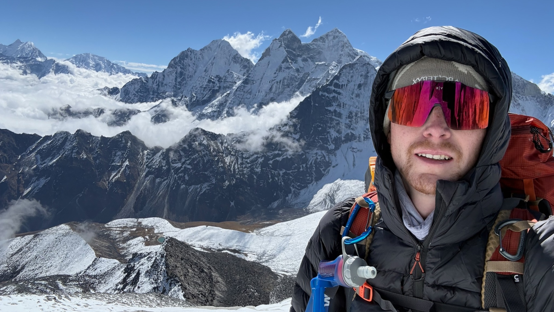This image shows a hiker wearing a puffy down jacket and mirrored ski goggles while standing atop a snow-covered mountain in the Everest region of Nepal.