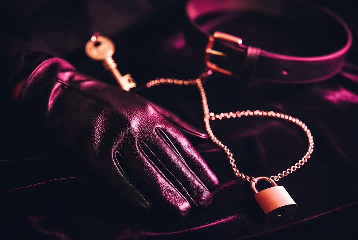 Black leather glove, golden key, and a chain with padlock beside a collar on dark velvet, symbolizing the dominant submissive dynamic