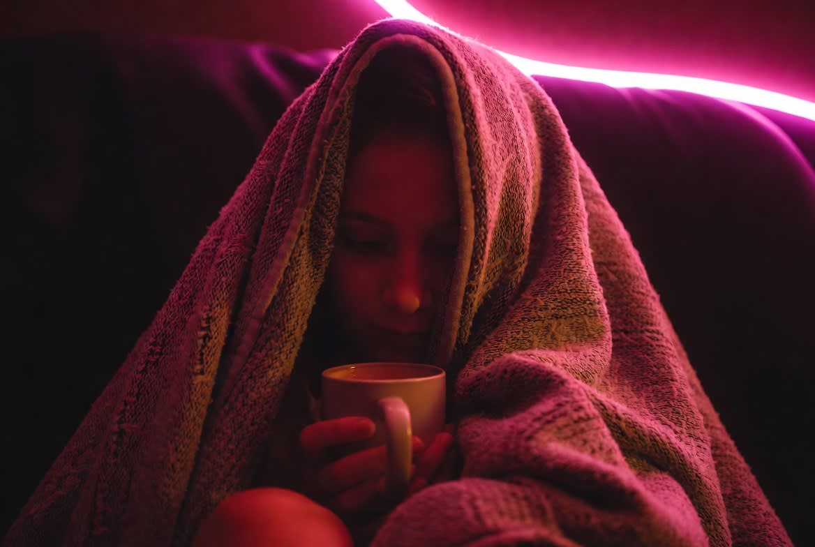 A person wrapped in a soft blanket holding a warm mug in dim pink light, a calm aftercare moment