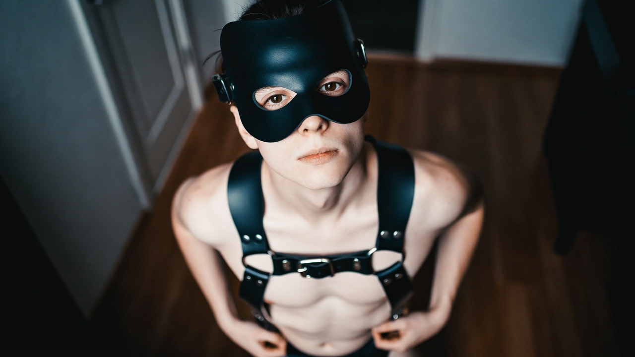 A masked submissive in a black leather harness kneels and gazes up at the camera, framed from above.