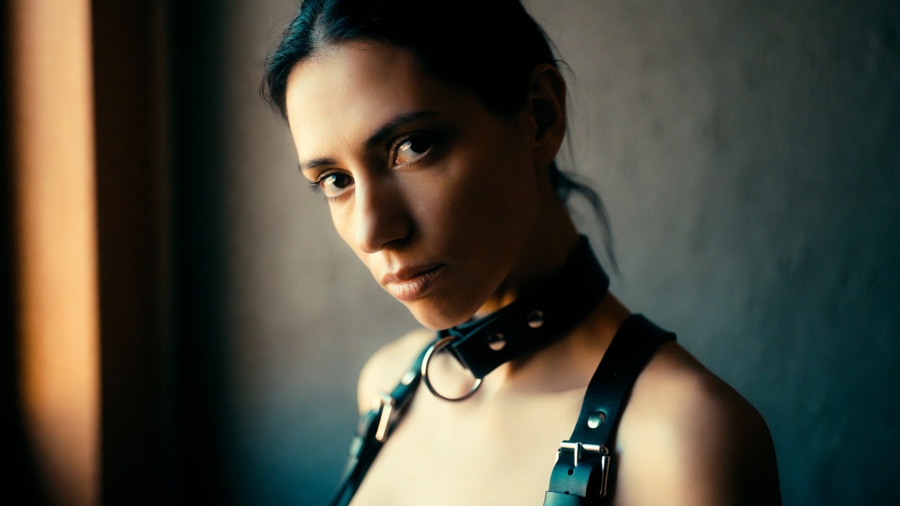 A woman in a black leather collar and harness, half her face in warm window light and half in cool shadow, holding a steady, unflinching gaze.