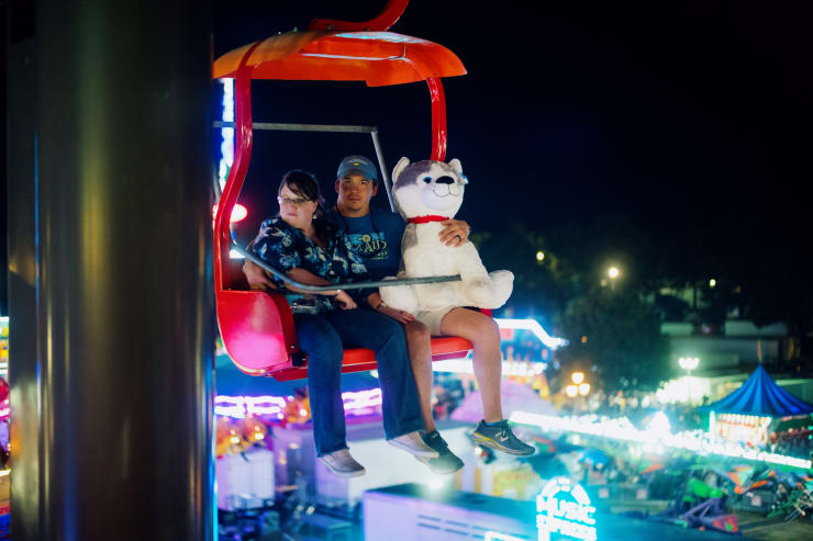 a woman and man with a large plush dog sitting in a ski lift above fair grounds at night