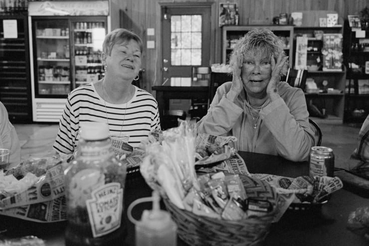 two older women are laughing at a table, the one on the right is making a funny face