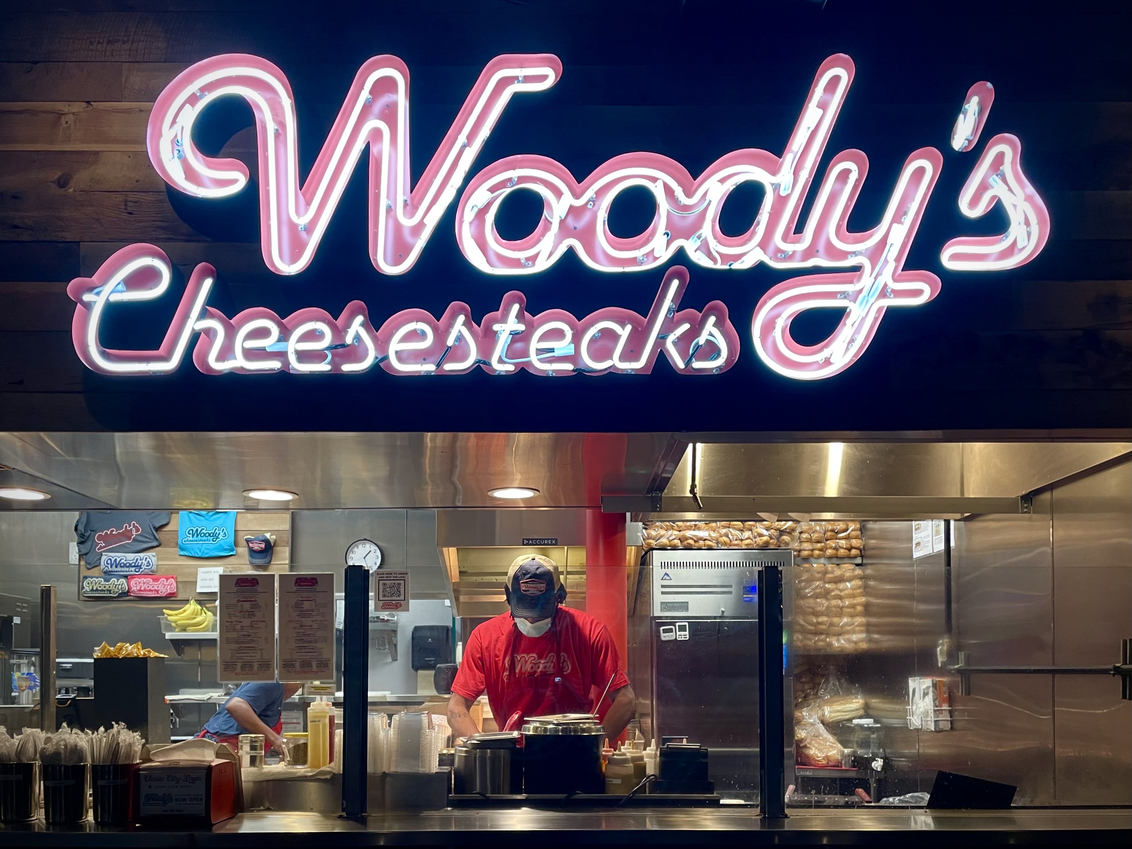 large neon sign reads Woody's Cheesesteaks above a kitchen where man and woman are preparing sandwiches