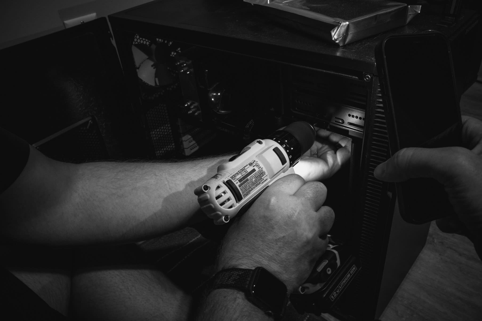 man using phone as flashlight while another man drills a hole to install a hard drive rack into computer case