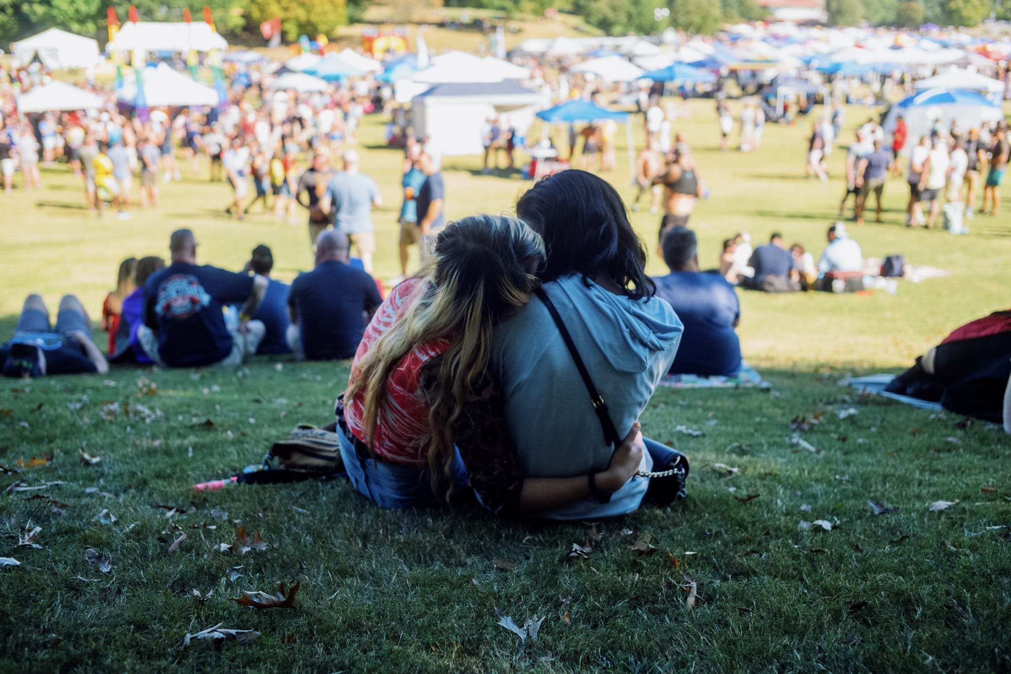 the back of two women hugging and kissing on the lawn of a park with a large crowd of people in the background