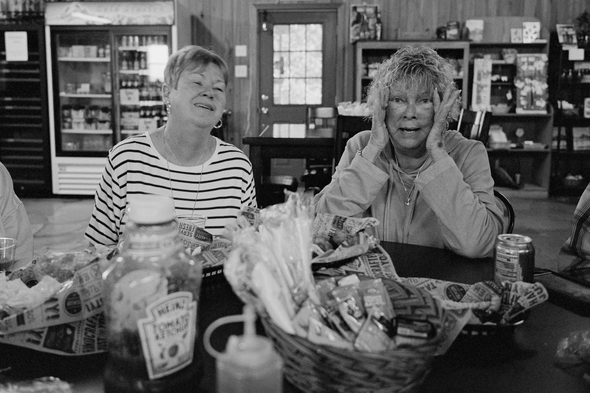 two older women are laughing at a table, the one on the right is making a funny face