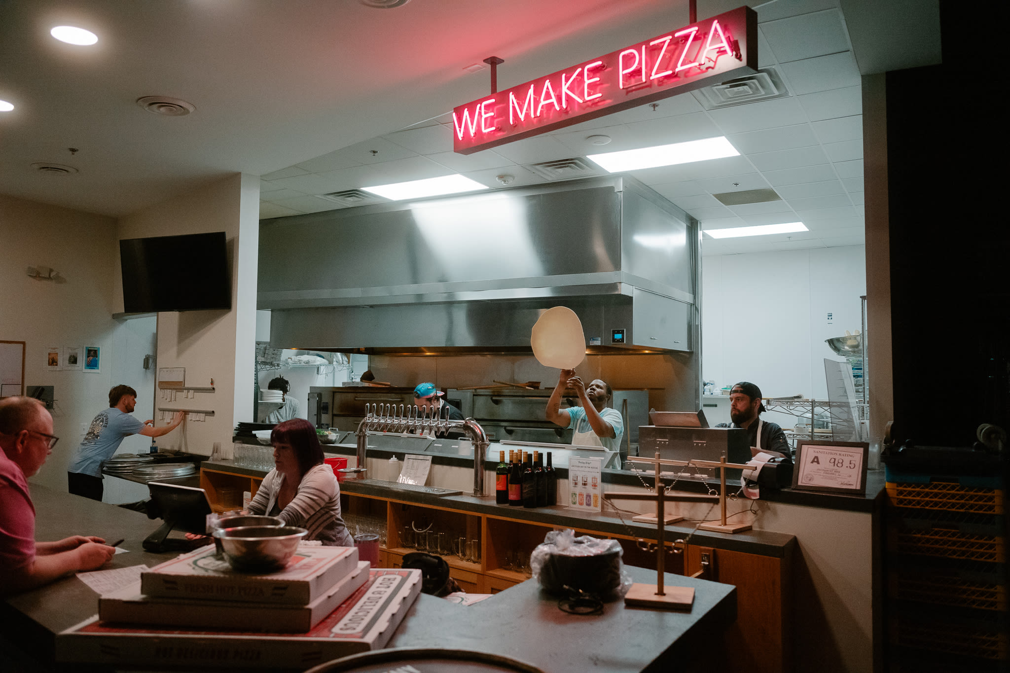 a man places an order at the counter of a pizza restaurant. there's a man tossing dough in the air under a neon sign that reads 'we make pizza'