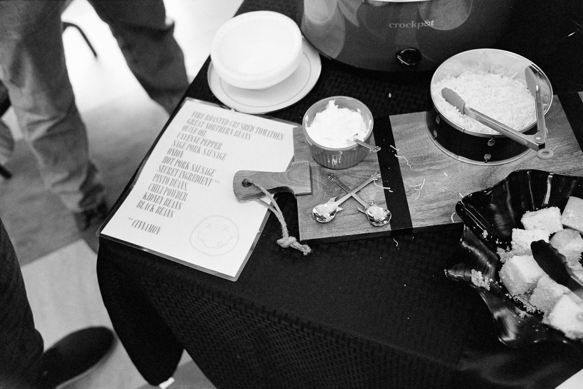 the corner of a table with bowls, a crockpot, chili toppings, cubes of cornbread and a list of ingredients