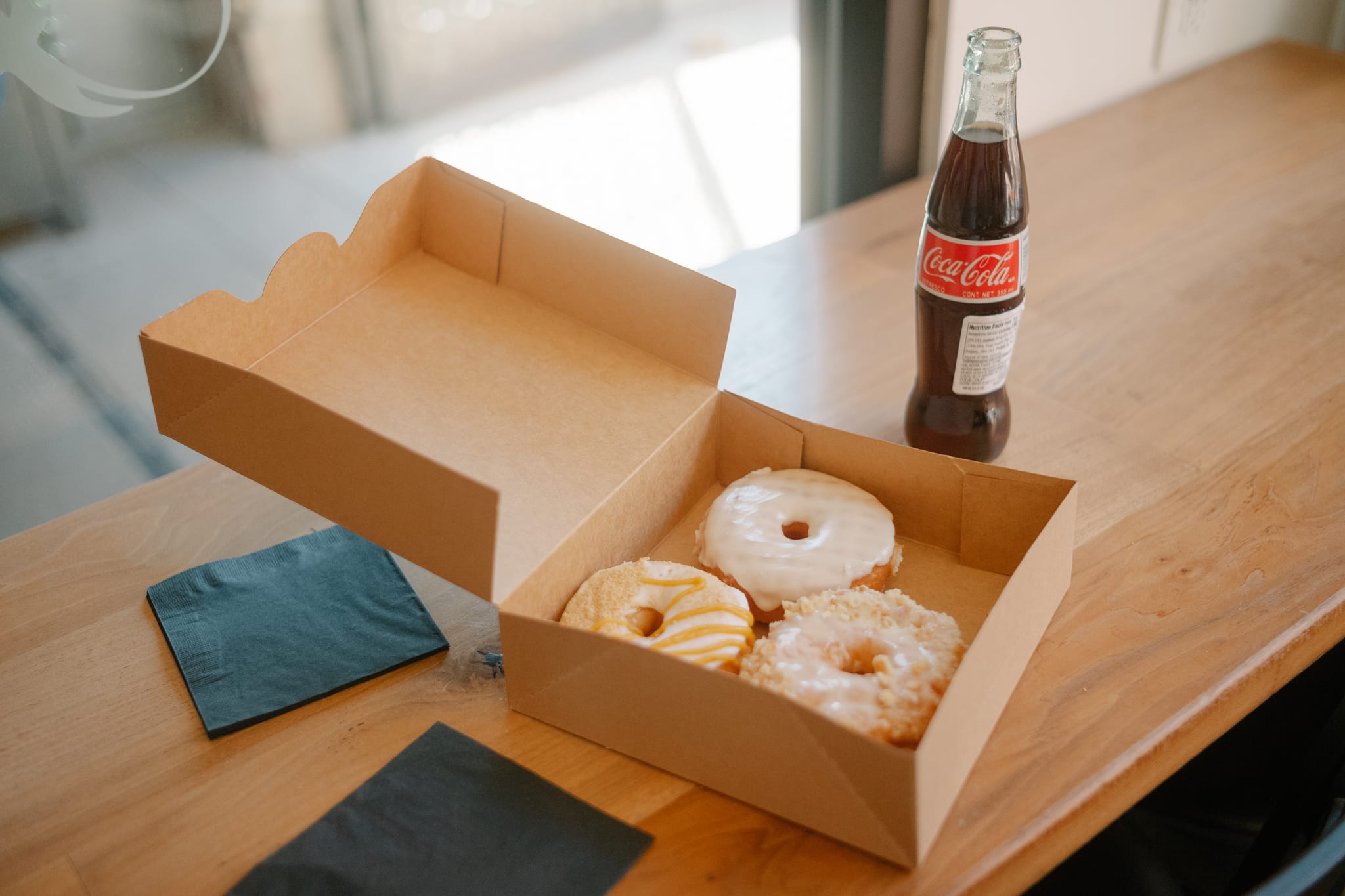 a small box of doughnuts sits on a wooden table next to a glass bottle of mexican coca cola