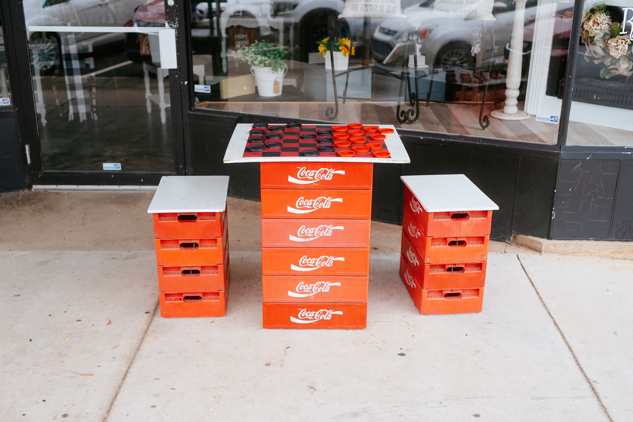 on a sidewalk sits an oversized checkers board sits on stacked coca cola crates with two stools made of smaller coca cola crates