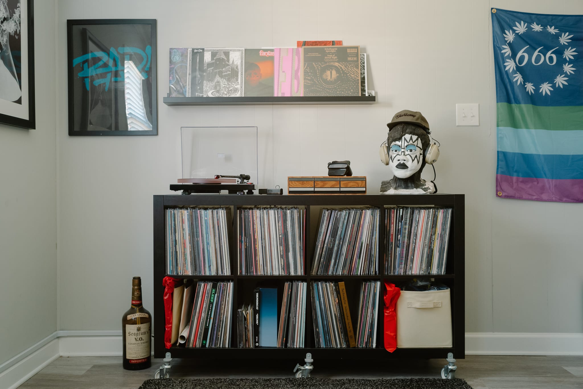 a standalone shelf of vinyl records with a turntable, cassette tape drawers and a bust of Lionel Richie painted like Ace Frehley from KISS sitting on top. another shelf mounted to the wall has various records on display