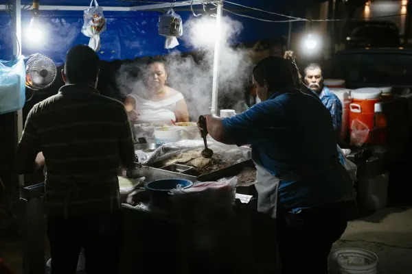 A taco street vendor cooking meat on a griddle and serving patrons