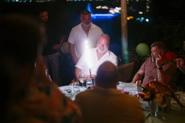 A man surrounded by friends, smiling at his birthday cake while a sparkler/candle is going off