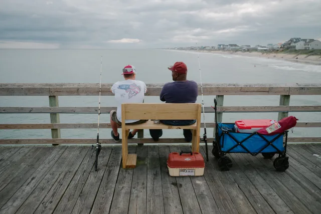 two men from behind sit on a bench fishing and talking together