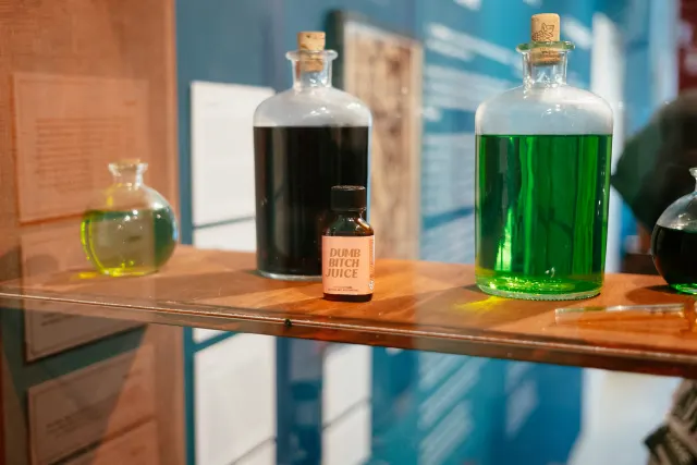 a shelf with various corked bottles and a small bottle of poppers that reads 'dumb bitch juice'