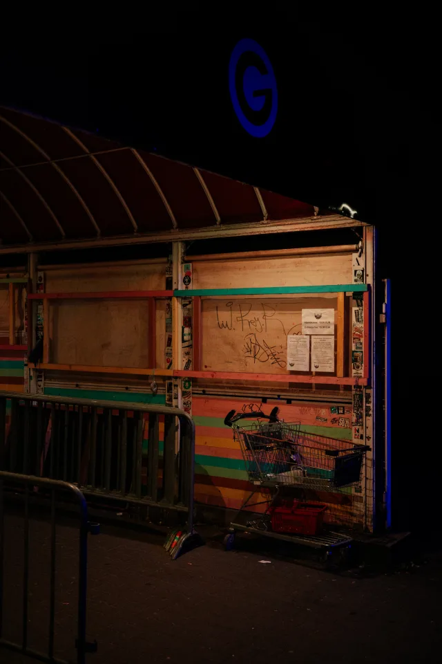 a grimy entrance to a club is dimly lit under an awning. a shopping cart with some bottles sits beside a wooden wall