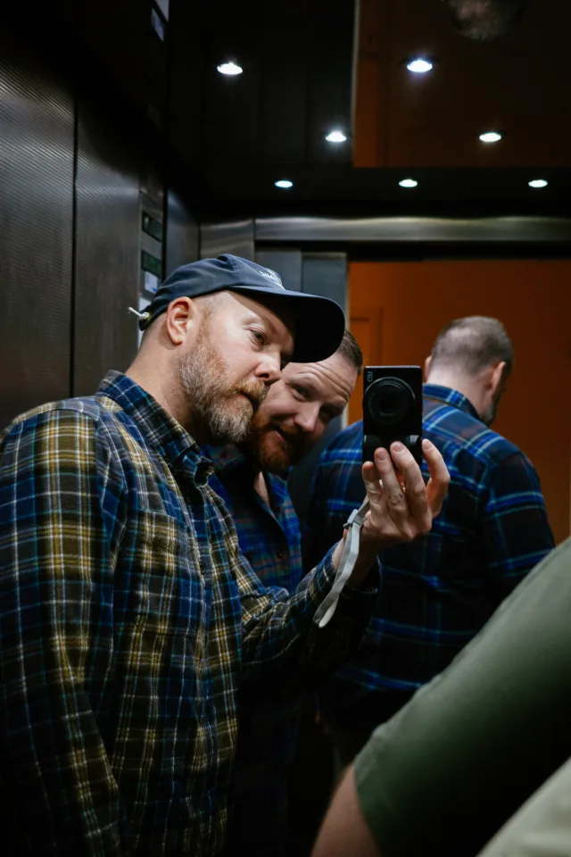four men in a mirrored elevator, one is pointing a camera at themselves