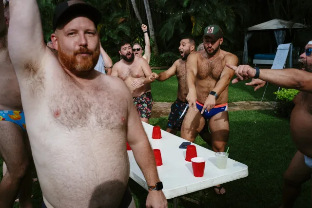 A group of men in swimsuits standing around a table, playing a drinking game. Half cheer in celebration, the other half protesting the outcome. One man in the foreground has his arms raised in victory