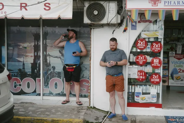 Two men stand outside of a pharmacy. The pharmacy heavily advertises things like HGH, Viagra, and pain killers.