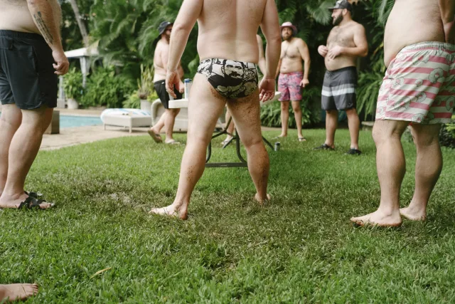 A bunch of men in swimsuits playing a drinking game. One's lower half is covered in mud.