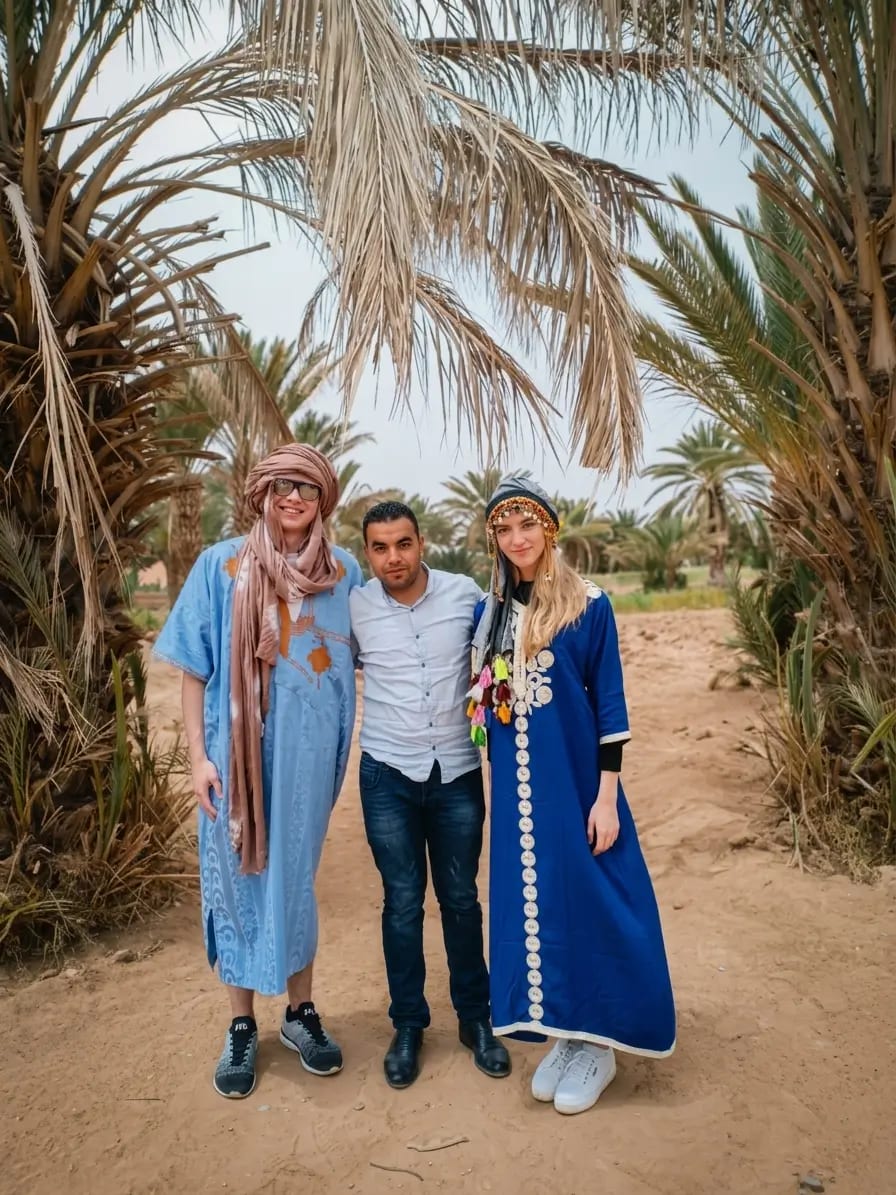 Hasy standing with two travelers in traditional dress at a palm-filled desert oasis