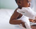 A toddler sits on an exam table as a gloved hand cleans her shoulder.