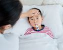 A mother feels the forehead of a young boy in bed with a thermometer in his mouth.
