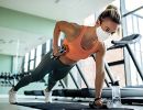 A woman wearing a mask in a gym does a one-armed plank with hand weights.