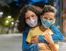 A woman hugs a child, both wearing face masks.