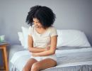 A woman sits on a bed, grimacing, with her arms wrapped around her torso.