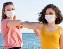 Two teenage girls wearing masks and summer attire bumping elbows.